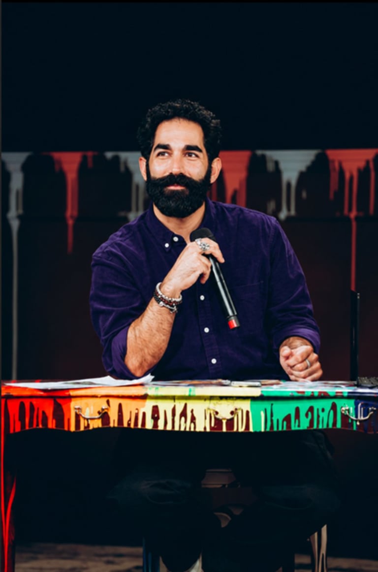 Man with dark beard holding microphone at panel table with colorful skyline backdrop